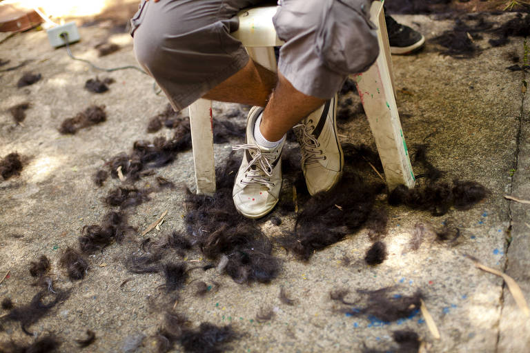 A imagem mostra uma pessoa sentada em uma cadeira plástica, com as pernas cruzadas sobre um chão de concreto. Abaixo, há uma quantidade significativa de cabelo escuro espalhado pelo chão, resultado de um corte de cabelo. A pessoa está usando um short cinza e tênis brancos. O ambiente é ao ar livre