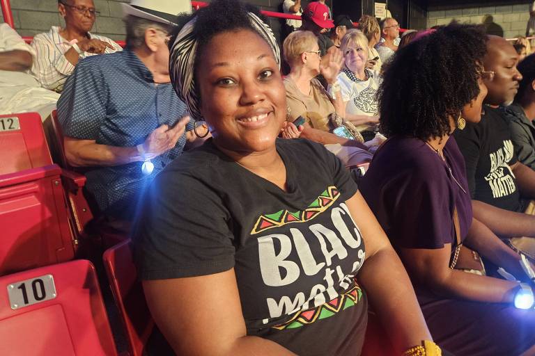 Uma mulher negra sorridente está sentada em uma cadeira em um evento, vestindo uma camiseta preta com a frase 'Black Lives Matter'. Ao fundo, há outras pessoas assistindo ao evento, algumas com expressões de interesse. A iluminação é suave e o ambiente parece ser um auditório ou teatro.