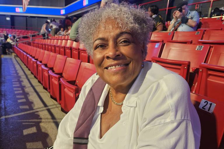 A imagem mostra uma mulher negra de cabelo grisalho, sorrindo enquanto está sentada em uma arquibancada. Ela usa uma blusa branca e parece estar em um ambiente de evento, com cadeiras vermelhas ao fundo e pessoas visíveis na distância.