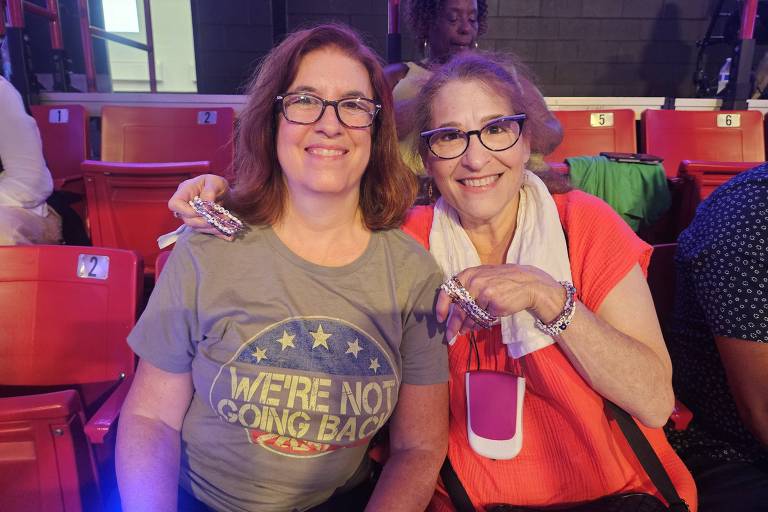 A imagem mostra duas mulheres sorrindo para a câmera em um ambiente de evento. A mulher à esquerda usa uma camiseta cinza com a frase 'WE'RE NOT GOING BACK' e a mulher à direita está vestindo uma blusa laranja. Ambas estão sentadas em cadeiras vermelhas, com um fundo que sugere um local de evento. Ao fundo, é possível ver outras pessoas e uma iluminação suave.