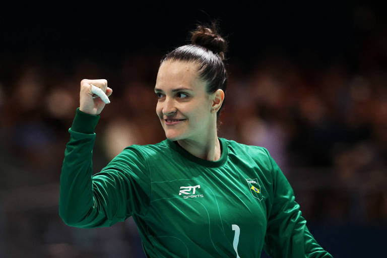 Mulher jogadora de handebol vestida com uniforme verde comemora com punho cerrado em ginásio coberto