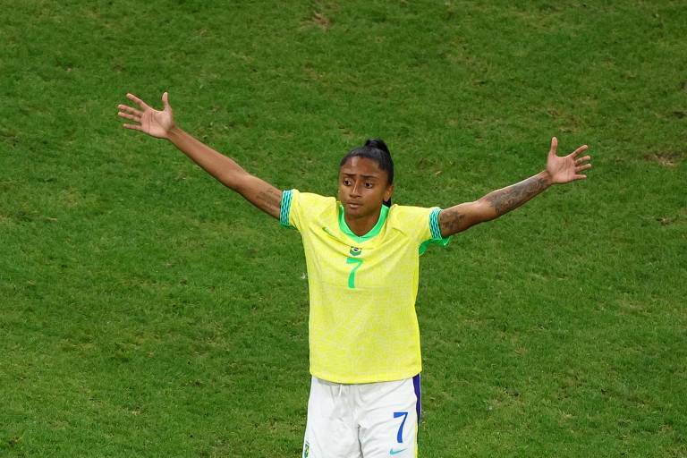 Uma jogadora de futebol está em campo, vestindo uma camisa amarela com detalhes verdes e um número 7. Ela está com os braços abertos, em uma pose expressiva, e parece estar se dirigindo a outros jogadores ou à torcida. O campo de futebol é visível ao fundo.