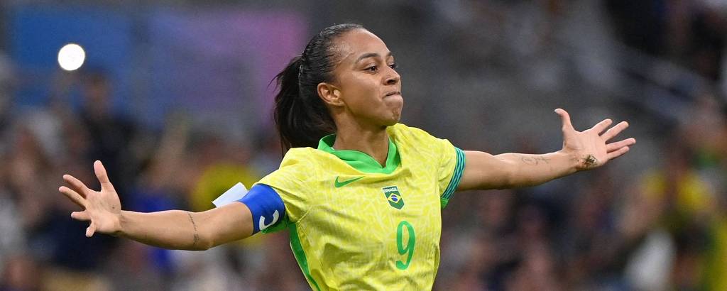 Uma jogadora de futebol vestindo uma camisa amarela com detalhes verdes e azuis, está comemorando com os braços abertos. O fundo mostra uma multidão em um estádio, sugerindo um ambiente de competição esportiva.