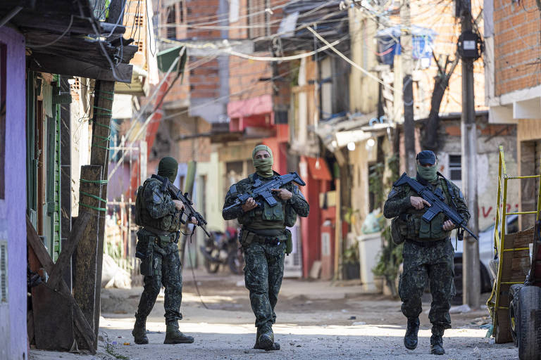 A imagem mostra três policiais armados patrulhando uma rua estreita. As casas ao redor são de alvenaria, com paredes coloridas e fios expostos.