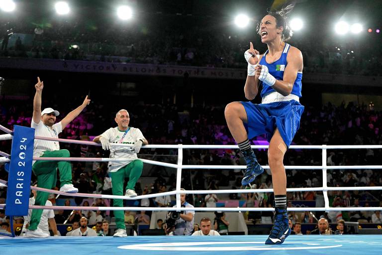 A imagem mostra um boxeador em um ringue, vestindo um uniforme azul. Ele está em uma pose de celebração, levantando um dedo em sinal de vitória. Ao fundo, duas pessoas, uma à esquerda e outra à direita, estão torcendo, vestindo roupas brancas e verdes. O ambiente é iluminado com luzes brilhantes, sugerindo um evento esportivo emocionante.
