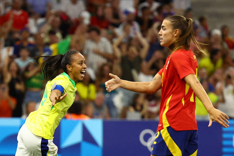 A imagem mostra duas jogadoras de futebol em um momento de interação durante uma partida. A jogadora à esquerda, vestindo uma camisa amarela com detalhes verdes, está comemorando com um sorriso, enquanto a jogadora à direita, vestindo uma camisa vermelha e calças escuras, parece estar se aproximando. Ao fundo, há uma multidão de torcedores, criando um ambiente animado.
