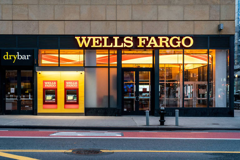 A imagem mostra a fachada de uma agência do Wells Fargo, com o nome 'WELLS FARGO' em letras grandes e iluminadas. À esquerda, há uma loja chamada 'drybar'. Na parte inferior da fachada, existem dois caixas eletrônicos com a marca Wells Fargo, em um fundo amarelo. A entrada da agência é visível, com portas de vidro. O ambiente parece ser urbano, com calçadas e sinalização de trânsito.
