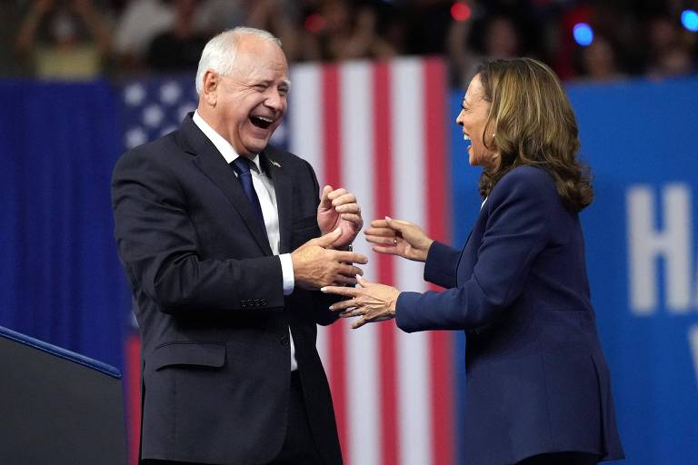 A imagem mostra Walz, um homem branco idoso de cabelos brancos, sorrindo próximo a Kamala, uma mulher negra de terno. Ao fundo, há uma bandeira dos Estados Unidos e um painel azul.