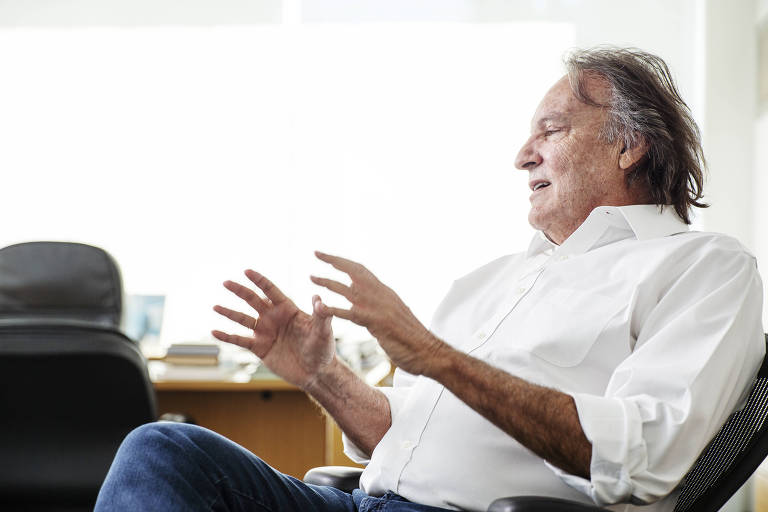 Um homem de cabelos grisalhos e pele clara está sentado em uma cadeira, gesticulando com as mãos enquanto fala. Ele usa uma camisa branca e jeans. Ao fundo, há uma mesa com alguns objetos e uma parede clara, iluminada por luz natural.
