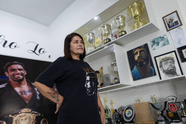 A imagem mostra uma mulher em pé em um ambiente decorado com prêmios e troféus. Ela é branca de cabelos lisos e está vestindo uma camiseta preta. Ao fundo, há prateleiras com vários troféus e quadros com retratos, incluindo uma grande imagem de um lutador