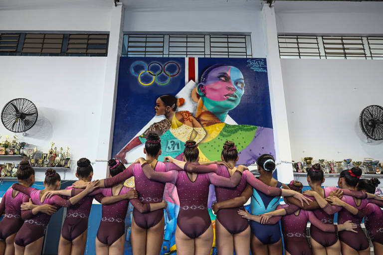 A imagem mostra um grupo de ginastas de costas, vestindo trajes de competição em tons de rosa. Elas estão com os braços entrelaçados, demonstrando união e apoio mútuo. Ao fundo, há um mural colorido que adiciona um ambiente vibrante à cena.