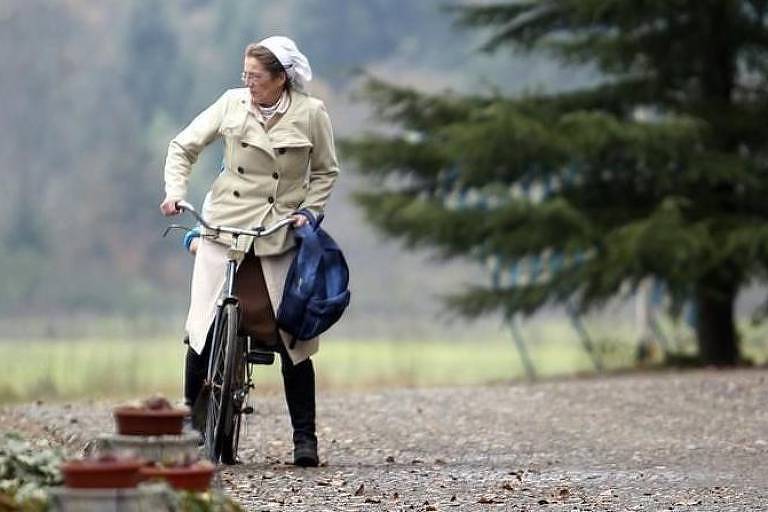 Uma mulher vestindo um casaco claro e uma faixa na cabeça está parada ao lado de uma bicicleta em um caminho de cascalho. Ela segura uma bolsa azul e olha para o lado, enquanto ao fundo há árvores e um campo desfocado.