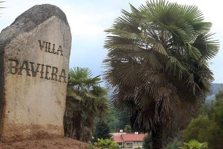 A imagem mostra uma placa de pedra com a inscrição 'VILA BAVIERA'. Ao fundo, há palmeiras e uma construção visível, com um céu nublado ao fundo.