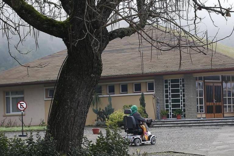 A imagem mostra uma pessoa sentada em uma cadeira de rodas motorizada, se deslocando em direção a um edifício de um andar. O edifício tem uma fachada simples, com janelas grandes e uma porta de vidro. À esquerda, há um grande árvore sem folhas e um sinal de proibição. O ambiente é nebuloso, sugerindo um clima frio e úmido.