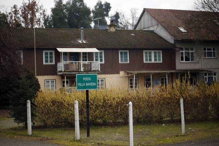 A imagem mostra um edifício de dois andares com uma fachada em tons de marrom e cinza. Na frente, há um sinal verde que diz 'POSTA VILLA BAVIERA'. O ambiente é cercado por uma cerca branca e há arbustos amarelos na frente. O céu está nublado e há árvores ao fundo.