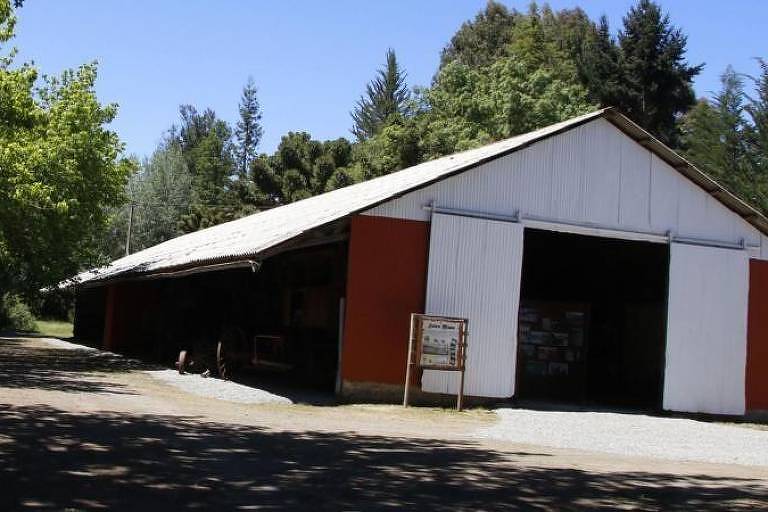 A imagem mostra um galpão de estrutura metálica com telhado de duas águas, pintado de branco e laranja. O galpão está localizado em uma área arborizada, com árvores verdes ao fundo e um caminho de cascalho à frente. Não há pessoas visíveis na imagem.