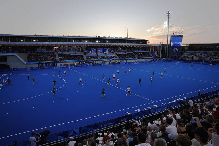 Uma partida de hóquei em campo está em andamento em um campo azul sob um céu ao entardecer. Jogadores de duas equipes estão em ação, com alguns usando uniformes brancos e outros em verde. O público está assistindo nas arquibancadas, que estão parcialmente ocupadas. Há uma tela grande visível ao fundo, mostrando a partida. A iluminação do campo é adequada para o jogo noturno.
