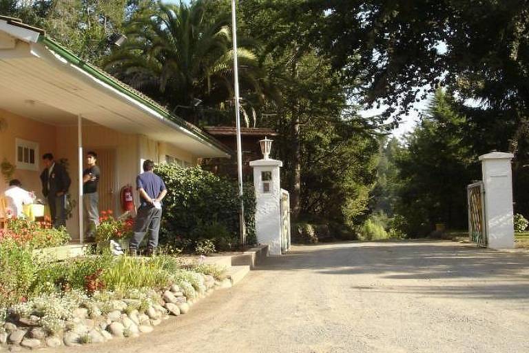 A imagem mostra a entrada de uma propriedade rural com uma casa de um andar à esquerda, cercada por um jardim com flores e plantas. Algumas pessoas estão na varanda e outras estão próximas à entrada. À direita, há um portão de entrada com colunas brancas e um caminho de terra que leva para dentro da propriedade, cercado por árvores e vegetação.