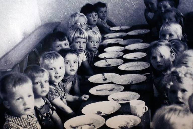 A imagem mostra um grupo de crianças sentadas em uma longa mesa, com pratos e copos à sua frente. As crianças estão olhando para a câmera, algumas com expressões curiosas. A mesa está cheia de pratos brancos e alguns copos. O ambiente parece ser uma sala de refeição, com paredes claras e um banco ao lado da mesa.