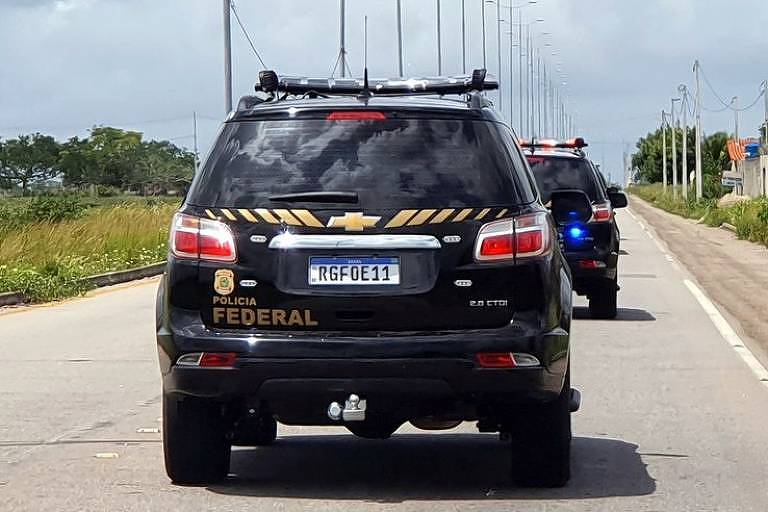 Imagem da parte trás de carro da Polícia Federal em uma estrada. O carro é preto e tem "Polícia Federal" escrito em amarelo na lataria. O dia está claro e o céu tem nuvens. 