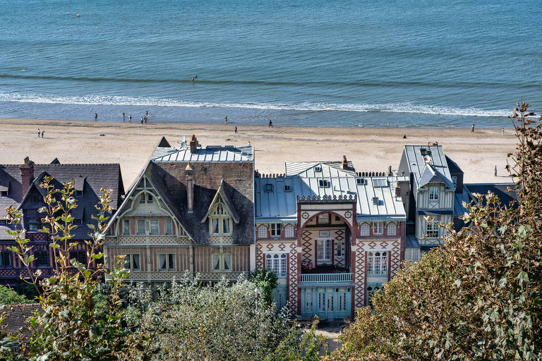 A imagem mostra uma vista aérea de um conjunto de edifícios próximos à praia. Os edifícios têm arquitetura variada, com telhados de diferentes estilos e cores. Ao fundo, é possível ver a areia da praia e o mar. Algumas pessoas estão caminhando na praia
