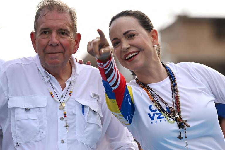 A imagem mostra duas pessoas em um evento. À esquerda, um homem de cabelo grisalho, vestindo uma camisa branca e colares coloridos, está sorrindo. À direita, uma mulher com cabelo preso, usando uma camiseta branca com detalhes coloridos e colares, está apontando para algo fora do quadro, também sorrindo.