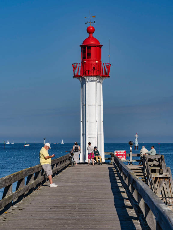 A imagem mostra um farol vermelho localizado em um pier de madeira que se estende sobre a água. Algumas pessoas estão presentes no pier, algumas sentadas e outras em pé, enquanto barcos podem ser vistos ao fundo