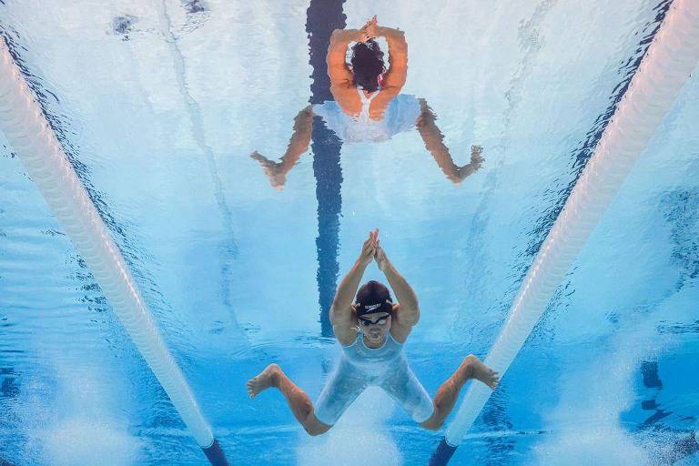 A imagem mostra uma nadadora submersa em uma piscina, com a cabeça para baixo e as mãos unidas acima da cabeça. A superfície da água reflete a imagem da nadadora, criando um efeito visual interessante. A nadadora está usando um traje de banho claro e um touca de natação escura. As raias da piscina são visíveis ao fundo, e a água é de um tom azul claro.
