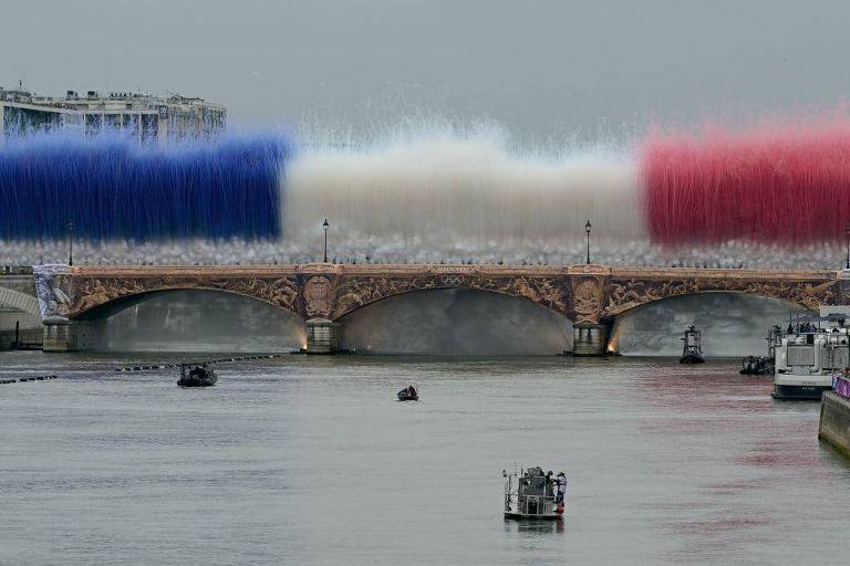 A imagem mostra uma ponte com uma grande explosão de cores, representando as cores da bandeira da França: azul, branco e vermelho. A explosão de cor se eleva acima da ponte, enquanto barcos navegam no rio abaixo. O céu está nublado, e a ponte apresenta detalhes ornamentais.
