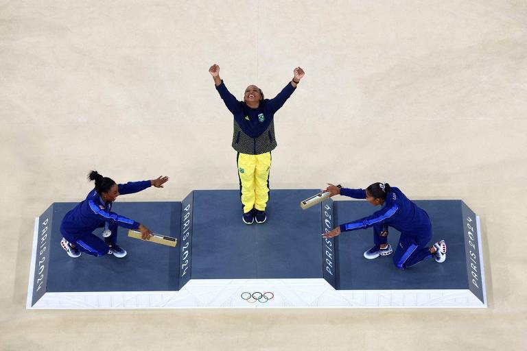 A imagem mostra uma cerimônia de premiação olímpica. No centro, uma atleta vestindo um uniforme amarelo e verde levanta os braços em celebração. À esquerda e à direita, duas atletas em uniformes azuis estão se preparando para entregar bastões. O cenário é marcado por um pódio com a inscrição 'PARIS 2024' e os anéis olímpicos na parte inferior.
