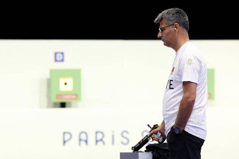 Um homem está em pé, segurando uma pistola, em um ambiente de competição de tiro. Ele usa uma camiseta branca e óculos de proteção. Ao fundo, há um painel com o texto 'PARIS' e alvos de tiro visíveis, incluindo um alvo verde com um ponto preto no centro. O homem parece concentrado e está posicionado ao lado de uma mesa.
