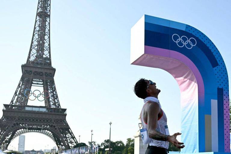 A imagem mostra um atleta em destaque, com uma expressão de esforço, em frente à Torre Eiffel. O atleta está usando uma camiseta sem mangas e um número de identificação visível em seu braço. Ao fundo, a Torre Eiffel se ergue sob um céu claro, e há um arco colorido com elementos olímpicos ao lado do atleta, simbolizando os Jogos Olímpicos. A cena transmite um clima de competição e celebração.
