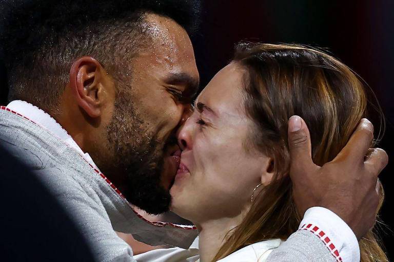 A imagem mostra um homem e uma mulher se abraçando e se beijando de forma carinhosa. O homem tem cabelo crespo e está vestindo uma jaqueta cinza com detalhes em vermelho. A mulher tem cabelo liso e castanho claro, e usa brincos. Ambos parecem emocionados e felizes, com expressões de alegria em seus rostos.
