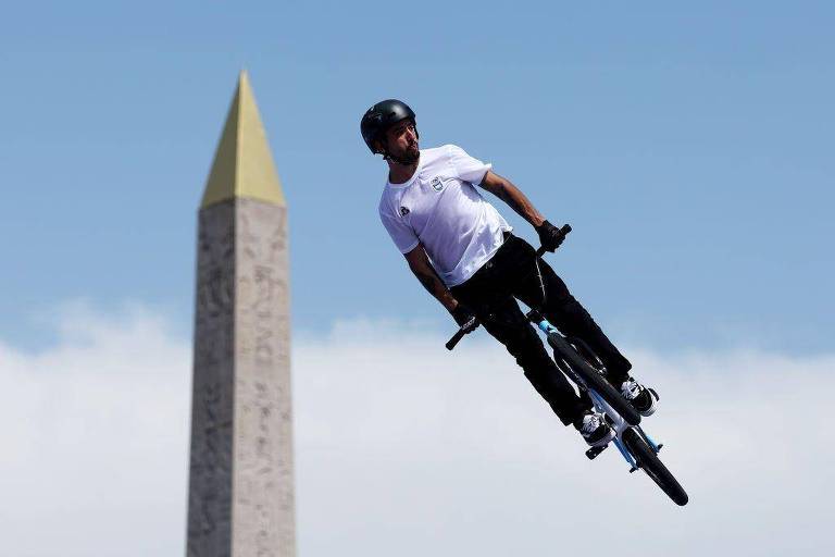 Um ciclista está realizando um salto acrobático em sua bicicleta BMX. Ele usa um capacete e uma camiseta branca, enquanto está no ar, com uma pirâmide de pedra ao fundo, que possui um topo dourado. O céu está claro e azul.
