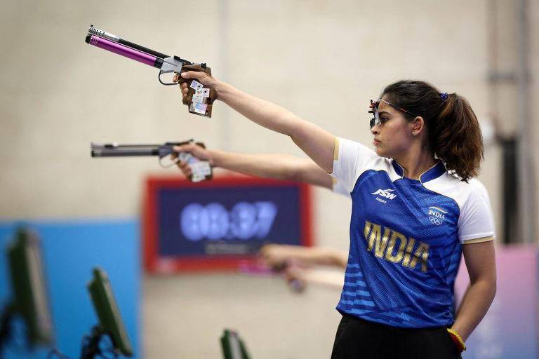 Uma atleta está competindo em uma prova de tiro com pistola. Ela está usando uma camiseta azul com detalhes brancos e a palavra 'INDIA' em letras douradas. A atleta está segurando duas pistolas, uma em cada mão, com a mão direita levantada e a mão esquerda estendida. Ao fundo, é possível ver um cronômetro marcando 08.37. O ambiente parece ser uma instalação de competição de tiro.
