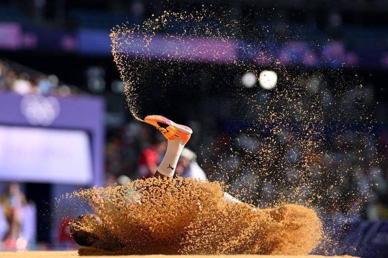 A imagem mostra uma atleta em um salto, caindo na areia. A perna da atleta está levantada, e a areia está sendo levantada em um spray ao redor. O fundo é desfocado, com uma multidão visível e um painel roxo ao fundo.
