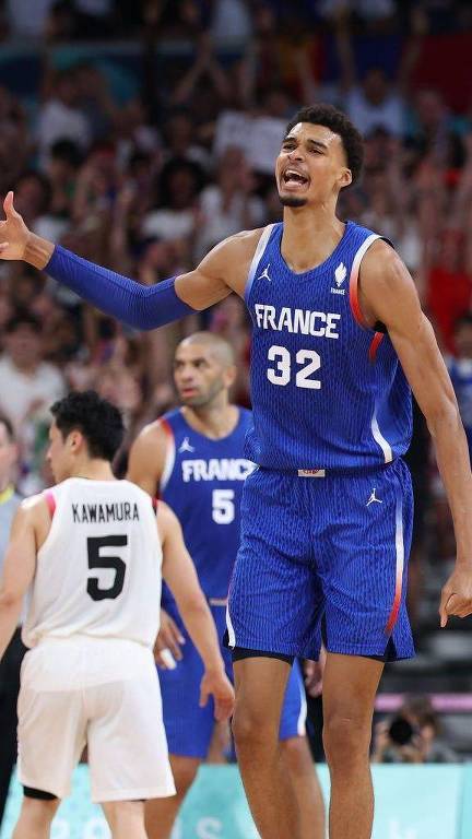 Um jogador de basquete, vestindo um uniforme azul com o nome 'FRANCE' e o número 32, está em uma posição expressiva, gesticulando com a mão direita enquanto parece estar reclamando ou se comunicando com os árbitros. Ao fundo, há uma multidão animada. Um jogador adversário, identificado como 'KAWAMURA' com o número 5, está de costas, olhando para o jogador da França. Outros jogadores estão visíveis no fundo.
