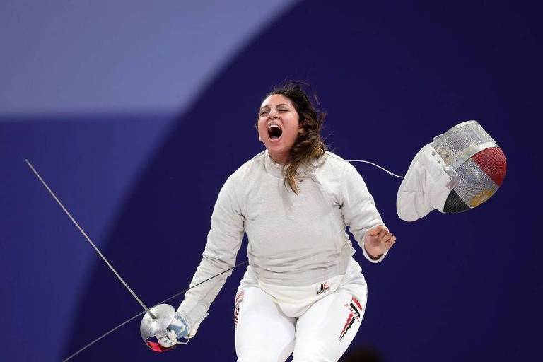 Uma atleta de esgrima está em uma pose de celebração, com um grito de alegria. Ela usa um uniforme de esgrima branco e segura uma espada na mão esquerda, enquanto a máscara de esgrima está pendurada em sua mão direita. O fundo é de um tom roxo, sugerindo um ambiente de competição.
