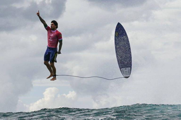 Um surfista está saltando sobre as ondas do mar, com uma mão levantada em sinal de vitória. Ele usa uma camiseta rosa e shorts azuis. A prancha de surf está presa a uma corda e se encontra em uma posição vertical, enquanto o céu está nublado ao fundo.
