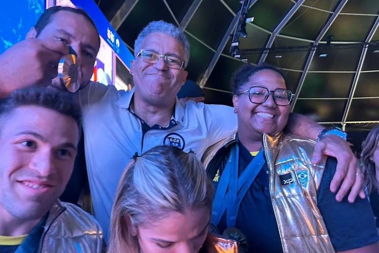 A imagem mostra um grupo de pessoas sorrindo em um ambiente festivo. Um homem à esquerda exibe uma medalha, enquanto outro homem ao centro sorri amplamente. Uma mulher à direita também sorri, usando um colete dourado. Ao fundo, há telas com imagens coloridas