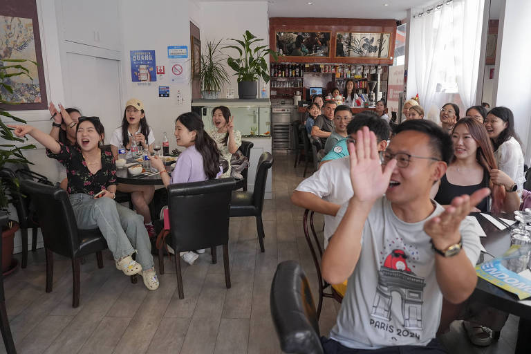 A imagem mostra um grupo de pessoas em um café, algumas delas estão rindo e se divertindo. À frente, uma mulher com blusa preta floral aponta para algo enquanto ri. Outras pessoas ao redor também estão sorrindo e interagindo. O ambiente é iluminado, com mesas e cadeiras pretas, e ao fundo há uma prateleira com bebidas e um aquário. As paredes têm algumas obras de arte e plantas decorativas.
