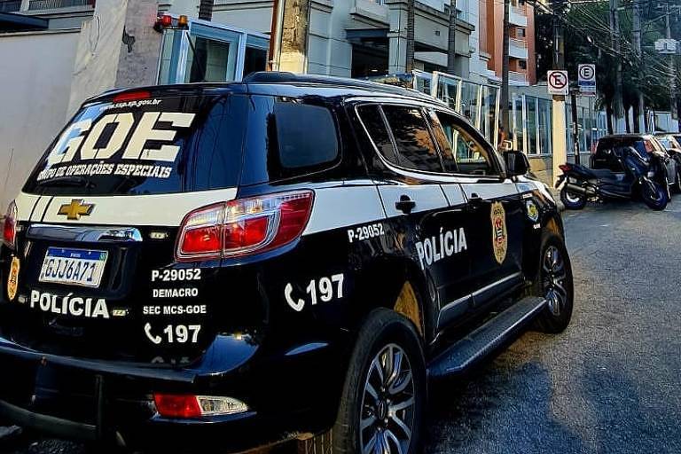 A imagem mostra uma viatura da polícia estacionada em uma rua. O veículo é preto e branco, com a inscrição 'GOE' (Grupo de Operações Especiais) em destaque. Há também a palavra 'POLÍCIA' e o número de telefone '197' visíveis. Ao fundo, pode-se ver prédios e algumas motos estacionadas