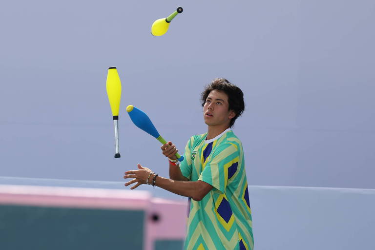 Um jovem está praticando malabarismo com três clavas coloridas, duas amarelas e uma azul. Ele usa uma camiseta com um padrão em zig-zag em tons de verde e amarelo. O fundo é simples e desfocado, destacando a ação do malabarismo.
