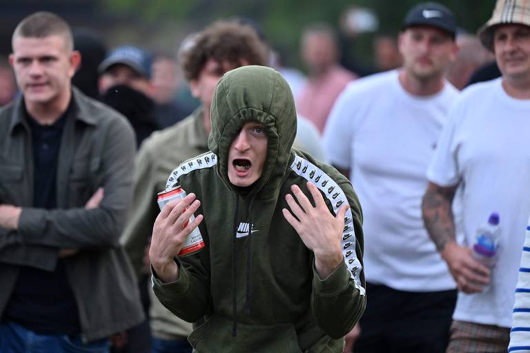 A imagem mostra um grupo de homens em um evento ao ar livre. Um homem em destaque, usando um moletom verde com capuz, está com as mãos levantadas e expressando uma emoção intensa. Ele segura uma lata de bebida na mão. Ao fundo, outros homens observam, alguns com expressões neutras e outros com sorrisos. O ambiente parece ser descontraído, com pessoas vestindo roupas casuais.