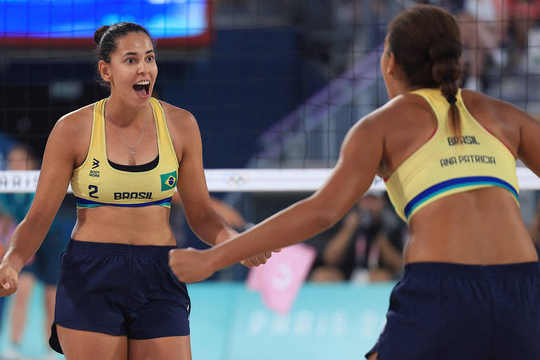 Duas jogadoras de vôlei de praia estão se celebrando em uma quadra. Ambas estão vestindo tops amarelos e shorts escuros, com expressões de alegria. O fundo mostra uma quadra de vôlei e uma plateia ao fundo.
