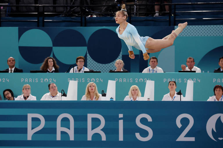 Uma ginasta está realizando um salto acrobático durante uma competição. Ela está vestindo um traje de ginástica azul claro e está no ar, com os braços estendidos e as pernas abertas. Abaixo dela, um painel com a inscrição 'PARIS 2024' e juízes observando a performance.
