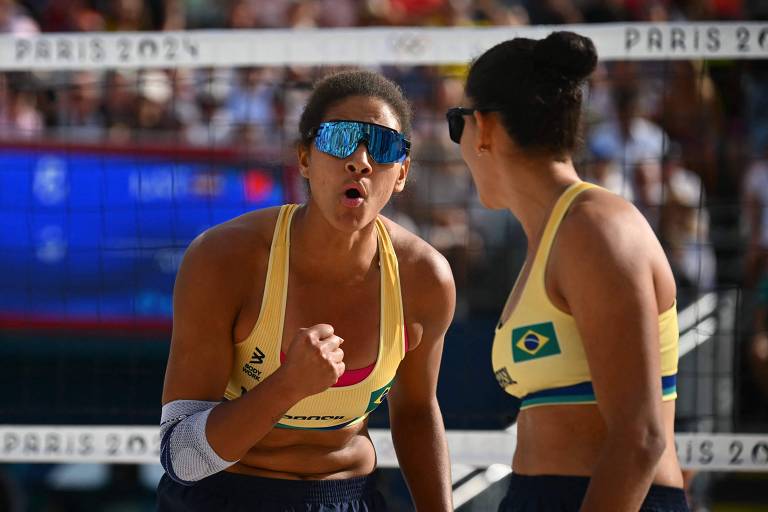 Duas jogadoras de vôlei de praia estão se animando durante uma partida. A jogadora à esquerda, com óculos escuros azuis, está gritando e fazendo um gesto de punho cerrado, demonstrando empolgação. Ambas estão vestindo tops amarelos com a bandeira do Brasil. Ao fundo, é possível ver a rede de vôlei e uma multidão assistindo ao jogo.
