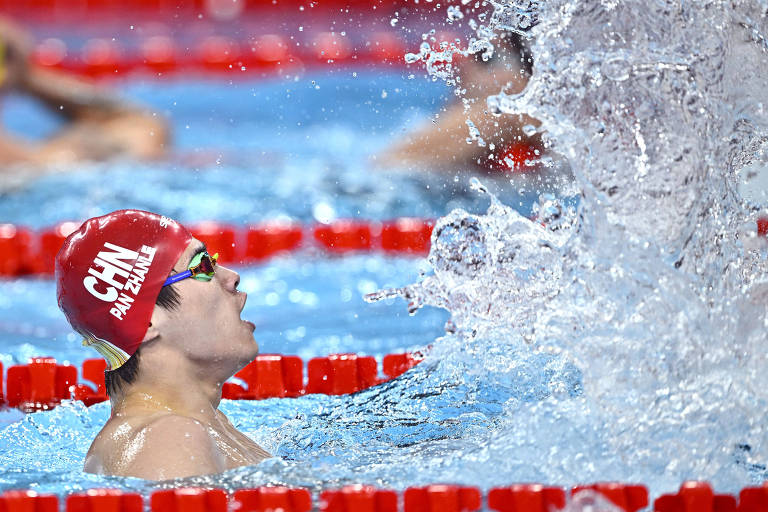 Um nadador com um boné vermelho e óculos de natação está em uma piscina, com água espirrando ao seu redor. O nadador parece estar se esforçando, com a boca aberta e a cabeça inclinada para trás. Ao fundo, é possível ver outras pessoas nadando.
