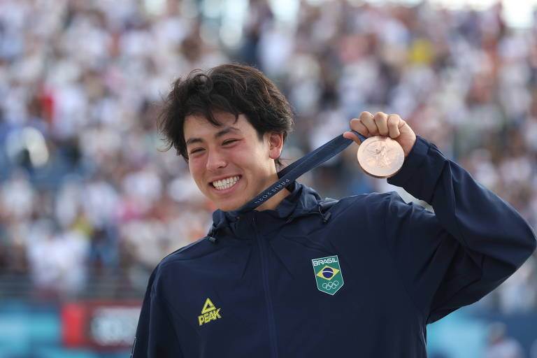 Um jovem atleta brasileiro sorri enquanto segura uma medalha de bronze com uma fita ao redor do pescoço. Ele está usando um agasalho azul com o logotipo da equipe brasileira e um emblema olímpico. Ao fundo, há uma multidão desfocada, sugerindo um evento esportivo.
