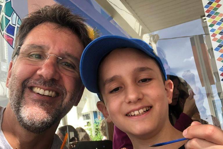 A imagem mostra um homem e um menino sorrindo enquanto seguram sorvetes. O homem, com cabelo escuro e barba, usa óculos e uma camiseta cinza. O menino, com um boné azul, segura um copo de sorvete de chocolate e uma colher, enquanto o homem segura um sorvete de baunilha em um cone. Ao fundo, há uma parede colorida e outras pessoas visíveis.